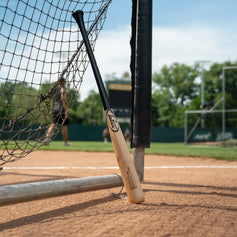 Tater X12 Pro balanced custom maple wood bat leaning against a batting cage net on a dirt infield