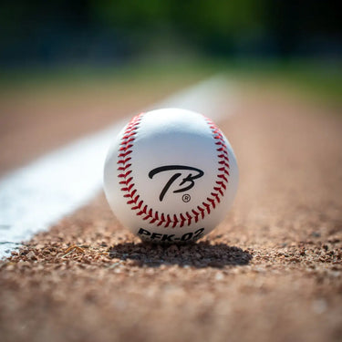 A white baseball with red stitching and a black stylized 'PB' logo, resting on a dirt baseball field near a white foul line.