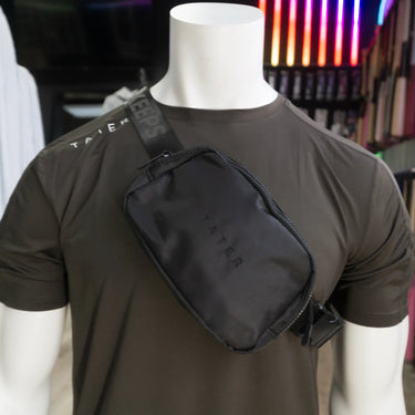 The image presents a mannequin dressed in an olive athletic T-shirt with a black crossbody fanny pack slung over the shoulder. The fanny pack features the Tater brand name embossed on the front, while the strap is adorned with the phrase "KEEPERS" in a repeating pattern, contributing to the sporty and stylish aesthetic of the product. The combination of the crossbody pack with the athletic wear suggests a trendy and practical accessory for sports enthusiasts or individuals with an active lifestyle. 