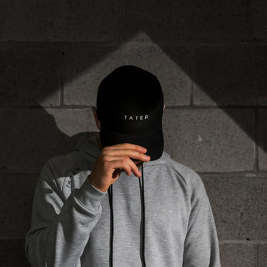 This image presents a person in a shadowy environment wearing a grey long-sleeve shirt and holding a black snapback cap with "TATER" embroidered in white across the front. The style is modern and minimalist, appealing to those who appreciate understated, sleek fashion. The cap is part of the Tater Baseball lifestyle collection, indicating a blend of sports and casual wear. The lighting and setting give the image an edgy, stylish vibe.