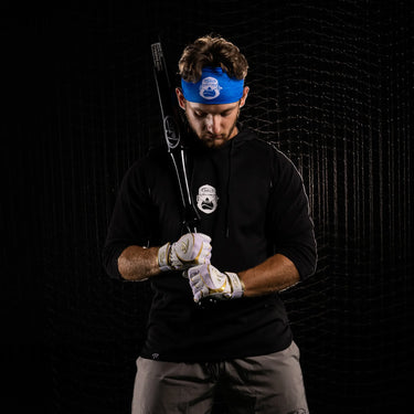 Baseball player adjusting batting gloves with bat, wearing blue headband and black hoodie in netted cage