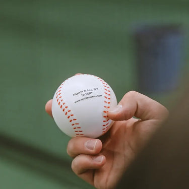 Hand holding white foam baseball with red stitching, labeled 'Foam Ball by Tater' for safe practice training.